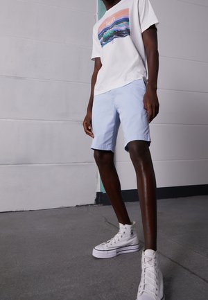 Mężczyzna w białym t-shircie z grafiką, jasnoniebieskich szortach i białych trampkach na wysokiej podeszwie stoi na betonowej podłodze przy szarej ścianie.