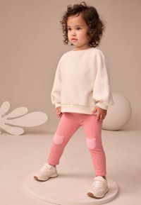 Hanorac crem din bumbac cu mâneci bufante, asortat cu colanți roz cu aplicații moi în formă de ursuleți pe genunchi. Adidași albi.