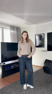 Jauna moteris stovi svetainėje vilkėdama smėlio spalvos megztinį, platėjančias tamsias džinsines kelnes ir baltus sportbačius su juodomis juostelėmis.