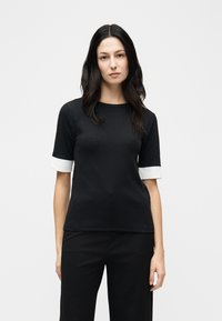 Γυναίκα με μακριά σκουρόχρωμα μαλλιά, ντυμένη με μαύρο ριγωτό μπλουζάκι με κοντά μανίκια και λευκά μανσέτα, καθώς και μαύρο παντελόνι, που στέκεται μπροστά σε λευκό ομοιόμορφο φόντο.