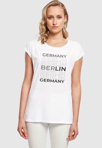 White t-shirt featuring "Germany" and "Berlin" text in varying fonts. Short rolled sleeves, relaxed fit, made from cotton.