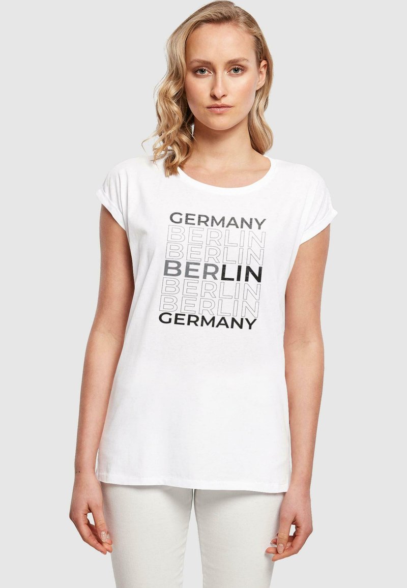 White t-shirt featuring "Germany" and "Berlin" text in varying fonts. Short rolled sleeves, relaxed fit, made from cotton.