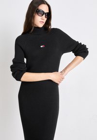 Svart ribbad stickad klänning med hög krage och långa ärmar. Har en liten flerfärgad logotyp på bröstet. Slät textur, figursydd silhuett.