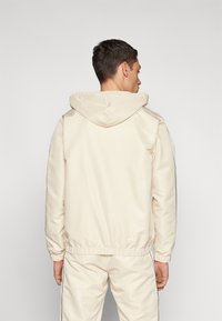 Sudadera crema con cordón en la capucha, puños elásticos y dobladillo. Textura suave, diseño ajustado y costuras sutiles a lo largo de los lados y la capucha.