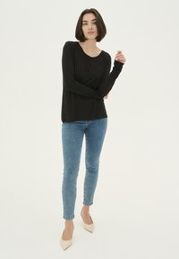 Fekete, hosszú ujjú felső, kerek nyakkivágással, kék farmernadrággal és bézs, hegyes orrú cipőkkel párosítva egy modellen.