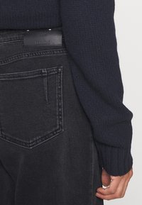 Personne portant un jean en denim noir et un pull texturé bleu marine foncé, se concentrant sur la poche arrière et la manche inférieure près de la main.