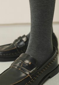 Čierne kožené mokasíny s dekoratívnym prešívaným detailom, doplnené o sivé bavlnené ponožky. Záber zblízka zdôrazňuje textúry a materiály.