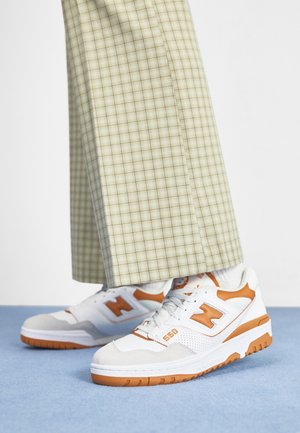 White and orange New Balance 550 sneakers worn with beige plaid wide-leg pants on a light blue floor.
