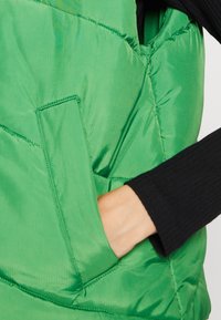 Zielona puchowa kamizelka z błyszczącym wykończeniem, z boczną kieszenią. Pod spodem częściowo widoczna czarna, prążkowana bluzka z długim rękawem.