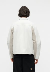 Άτομο με σκούρα κυματιστά μαλλιά φοράει λευκό μπουφάν με μπεζ μανίκια και μαύρο παντελόνι, με θέα από πίσω μπροστά σε απλό φόντο.