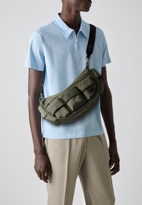 Sac en nylon vert olive à bandoulière avec plusieurs poches zippées, porté en travers du corps. Le modèle porte une chemise polo bleu clair et un pantalon beige.