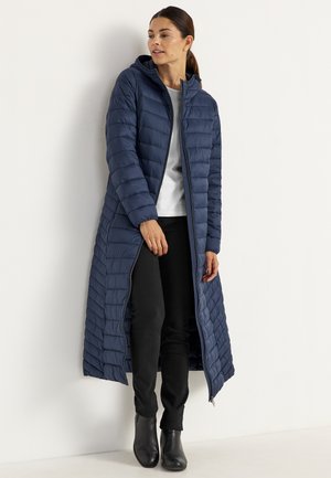 Lang marineblauwe pufferjas met horizontaal gestikte patroon, voorzijde met rits en capuchon. Gedeeltelijk gedragen over een witte top en zwarte broek, knielange uitstraling.
