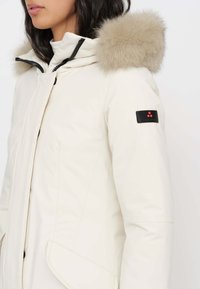 Giacca invernale beige chiaro con cappuccio foderato in pelliccia, dettagli neri e chiusura frontale con zip. Presenta una toppa sulla manica e dettagli testurizzati.