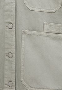 Primer plano de una tela de mezclilla beige claro con un gran bolsillo y un bolsillo más pequeño encima, junto a botones de presión metálicos en una prenda.