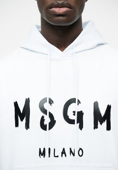 Κοντινό ενός ατόμου που φοράει λευκή κουκούλα με μαύρο κείμενο "MSGM" και "MILANO" στο μπροστινό μέρος.