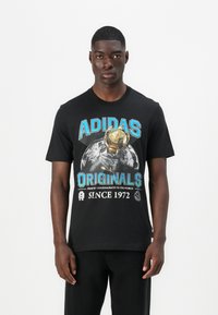 Fekete pamut póló, amelyen egy trófea és a Föld grafikus nyomata látható, kék és fehér színű "ADIDAS ORIGINALS" és "SINCE 1972" felirattal.