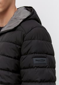 Schwarze Pufferjacke mit einer grauen Kapuzenfutter, mit einer gesteppten Textur und einem Gummilogo-Patch auf der linken Schulter.