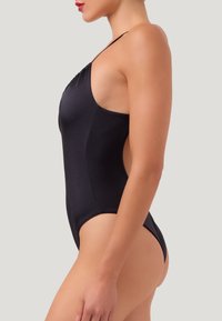 Persona snella in costume intero nero con spallina sottile, mostrata di lato su uno sfondo uniforme.