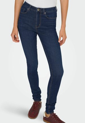 Tamsiai mėlyni siaurieji džinsai su kontrastinga oranžine siuvimu, penkios kišenės ir mygtukų užsegimas. Derinami su tamsiai raudonais sportbačiais.