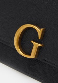 Zlaté kovové písmeno "G" připevněné na texturovaném černém koženém povrchu, pravděpodobně součást tašky nebo doplňku.