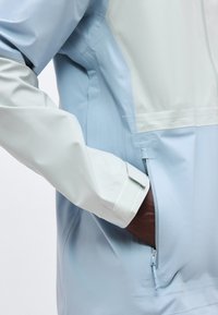 Κοντινό ενός χεριού που είναι μερικώς εισαγμένο στην τσέπη με φερμουάρ ενός ανοιχτό μπλε και λευκού μπουφάν με μανσέτα βέλκρο.