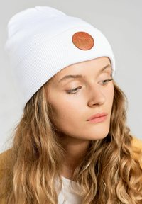 Biały prążkowany beanie z okrągłą brązową skórzaną łatką z motywem gór. Materiał ma teksturowany wygląd i dopasowany krój.