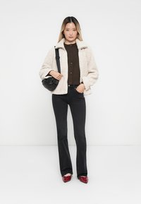 Veste en fausse fourrure crème, pull marron à boutons, jean noir évasé, sac à bandoulière noir et chaussures rouges. Fond simple avec une tonalité neutre.