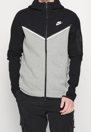 Osoba ubrana w czarno-szarą bluzę Nike na suwak z wysokim kołnierzem oraz czarne spodnie bojówki, ręce częściowo w kieszeniach.