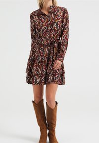 Abito a maniche lunghe in un motivo floreale rosso scuro con dettagli bianchi e verdi; vestibilità ampia, orlo arricciato, abbinato a stivali marroni sopra il ginocchio.