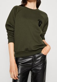 Donna che indossa un felpa verde oliva con la scritta "Athletic" e pantaloni di pelle nera, una mano in tasca, vista ritagliata dal collo ai fianchi.