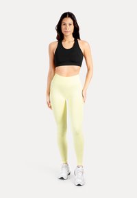 Zwarte sportbeha en lichtgele, hoog uitgesneden legging met een gladde textuur en strakke pasvorm, gecombineerd met witte sportschoenen.