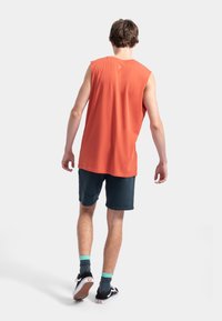 T-shirt en coton orange sans manches avec un ourlet arrondi, associé à un short sombre et des baskets noires. Présente un col rond et un design minimaliste.
