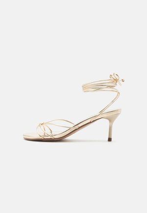 Zlaté sandále na remienky s nízkym ihličkovým podpätkom a okolo členka previazanými remienkami do mašle na bielom pozadí.