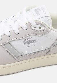 Sneakers Lacoste blancs et beiges avec des lacets blancs, panneaux en cuir texturé et logo crocodile Lacoste avec "EST. 1933" sur le côté.
