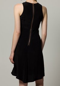 Svart ärmlös klänning med ett åtsittande liv och utsvängd kjol. Har en synlig koppardragkedja på baksidan och en slät tygtextur.