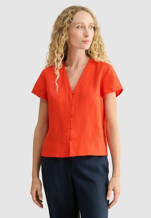 Mujer con cabello rubio rizado lleva una blusa corta de manga corta naranja brillante con botones y pantalones oscuros, mirando ligeramente hacia su izquierda.