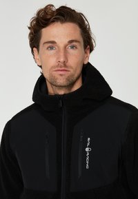 Zwarte fleecejack met een textuurkap, voorzien van gladde zwarte panelen, ritssluitingen en het witte "Sail Racing" logo aan de linkerkant.