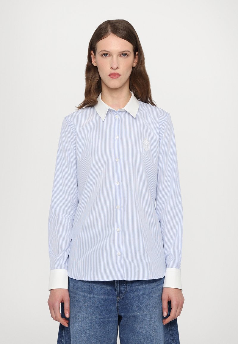 Light blue striped button-up shirt with white collar and cuffs, featuring a subtle emblem on the left side. Cotton fabric, tailored fit.