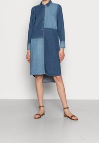 Sukienka koszulowa z denimu w różnych odcieniach niebieskiego, z paskiem na guziki, długimi rękawami i zaokrąglonym dołem. Połączona z brązowymi sandałami na ramiączkach.