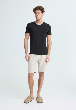 Noor mees seisab, seljas must lühikeste varrukatega V-kaelusega särk, heledat beeži tooni lühikesed püksid ja beežid libisemisjalatsid, taust on hele ja lihtne.