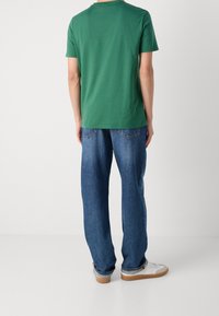 Vihreä puuvillapaita lyhyillä hihoilla yhdistettynä löysiin sinisiin farkkuihin ja valkoisiin lenkkareihin, näkyvillä saumoilla ja klassisilla suunnittelutiedoilla.