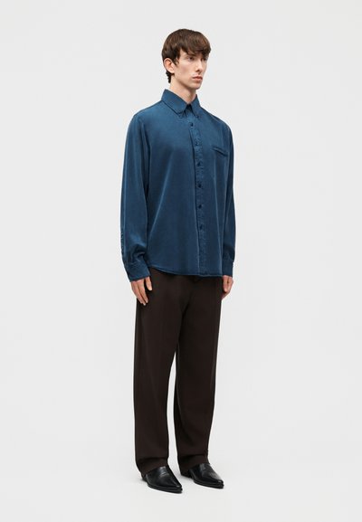 Νέος άνδρας που στέκεται, φοράει σκούρο μπλε μακρυμάνικο πουκάμισο με κουμπιά, άνετο μαύρο παντελόνι και μαύρα μυτερά παπούτσια σε ανοιχτό φόντο.