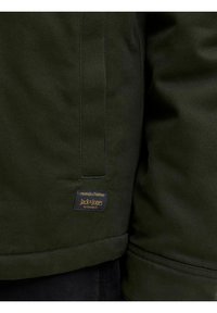 Veste en tissu vert foncé avec des coutures visibles et une petite étiquette noire portant la mention "Jack & Jones" près du bord inférieur côté gauche.