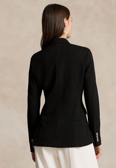 Femeie cu păr drept castaniu, îmbrăcată într-un sacou negru ajustat și pantaloni albi, văzută din spate pe un fundal bej.