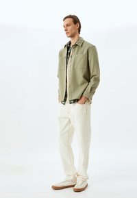 Camicia button-up verde chiaro con due tasche sul petto, indossata sopra una camicia a quadri, abbinata a pantaloni color crema e sneakers beige.