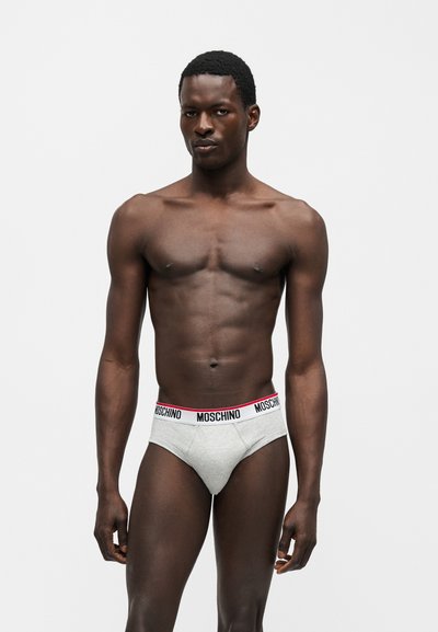 Άνδρας χωρίς πουκάμισο στέκεται μπροστά σε απλό φόντο φορώντας γκρι εσώρουχα με λευκή μέση που αναγράφει "MOSCHINO" με μαύρα γράμματα.