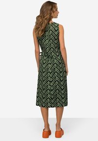 Grünes Kleid mit schwarzem Zickzackmuster, ärmelloses Design, tailliert, knielang, kombiniert mit orangen Plateau-Schuhen.