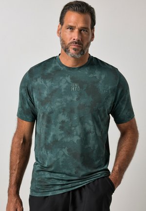Uomo con la barba che indossa una maglietta a maniche corte mimetica verde scuro con testo "UML MTD", in piedi con una mano in tasca.