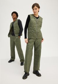 Ensemble en vert olive matelassé composé d'un gilet et d'un pantalon au design rembourré, avec une fermeture éclair, présenté sur un modèle portant des chaussures noires.