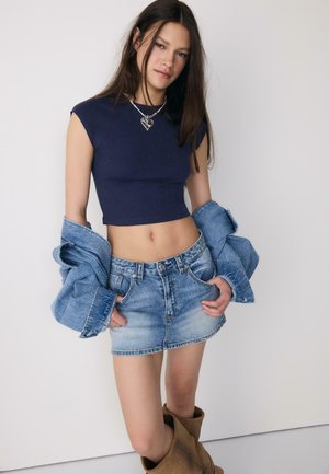 Mladá žena v námořnicky modrém krátkém topu, džínové mini sukni, držící džínovou bundu staženou z ramen a v hnědých vysokých kozačkách, stojící před prostou zdí.